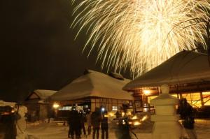 夜の大内宿雪まつりの写真002（雪をかぶった家の後ろに花火が上がるのを見る観光客）