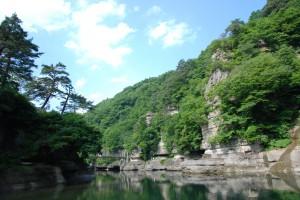 夏の塔のへつりの写真002（川から見上げる岩肌の上のほうは緑の木で覆われている）
